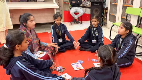 Un petit groupe d'étudiants assis en méditation dans un cercle avec une enseignante, dans le cadre des activités du défi de la santé mentale. Le cadre est informel et interactif, avec du matériel pédagogique visible sur le sol, ce qui témoigne d'un environnement d'apprentissage actif.