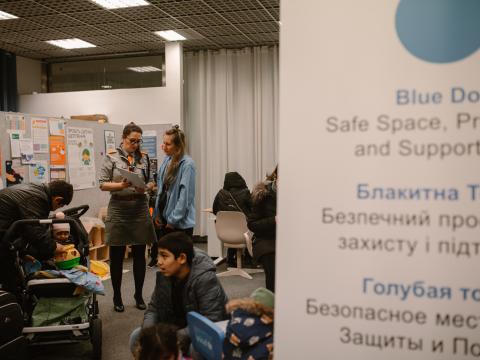 Two women talk in a room surrounded by adults and children sitting down.