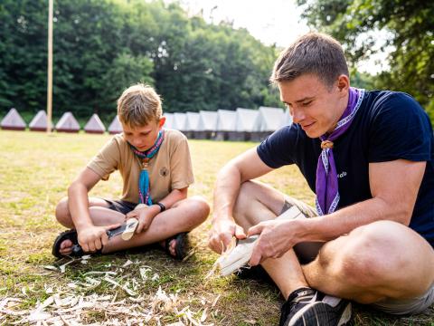 A scout leader and a Scout cut wood while sitting on the floor.