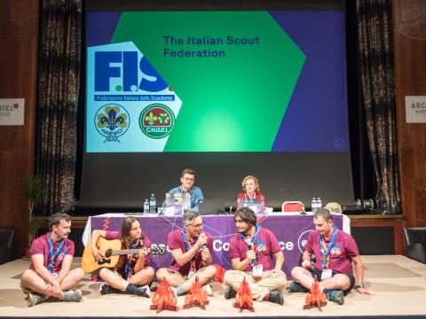 Scouts sit on a stage with small campfires made of cardboard on the floor.