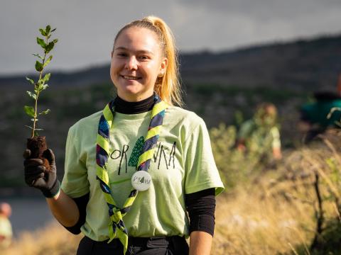 A girl wearing a shirt and a scarf holds a plant while smiling at the camera.