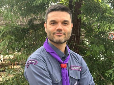 A man smiles at the camera. He wears a Scout uniform with a purple Scout scarf.