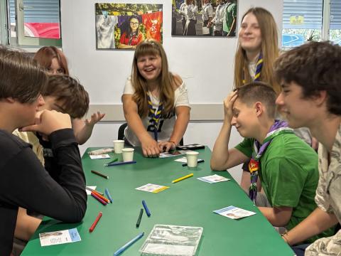 Seven young Ukrainians wearing Scout scarves sit around a table, some of them laughing.
