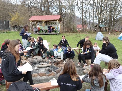 Young people of different ages sit around a campfire while holding papers.