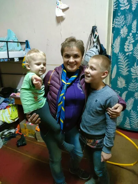 A Scout volunteer holds two Ukrainian children who are smiling after receiving new shoes from Spanish Scouts