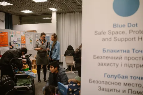 Two women talk in a room surrounded by adults and children sitting down.