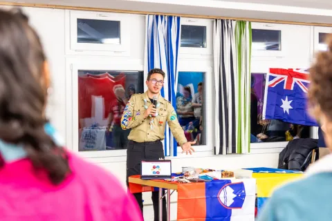 A man wearing a Scout uniform talks behind a desk with the Slovenian flag.