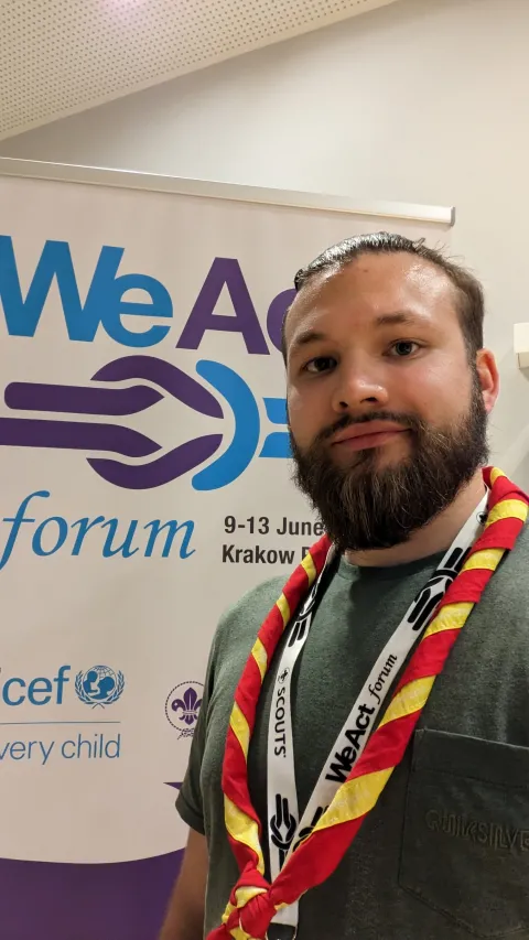 A man wearing a Scout scarf takes a selfie with a roll-up that reads 'WeAct Forum'.