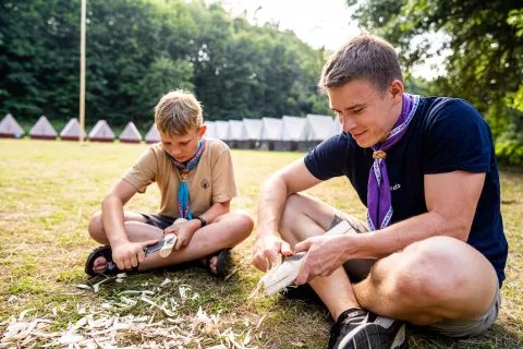A scout leader and a Scout cut wood while sitting on the floor.