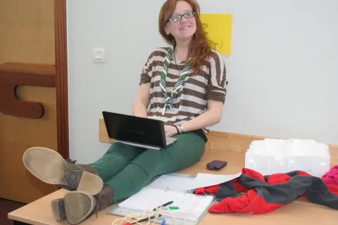 Máire seats on a table while using a computer and smiles.