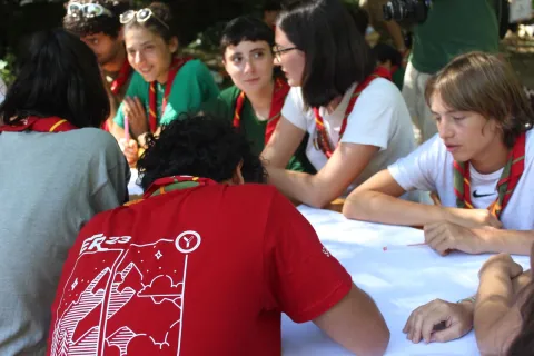 A group of young people write on a table. One of them is wearing a red t-shirt.