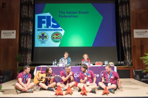 Scouts sit on a stage with small campfires made of cardboard on the floor.