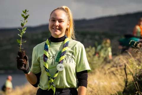 A girl wearing a shirt and a scarf holds a plant while smiling at the camera.