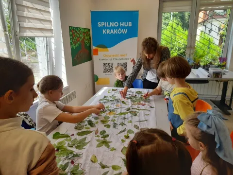 Ukrainian children place leaves on a table guided by an adult. On the wall there's a banner with the title "Spilno Hub Kraków".