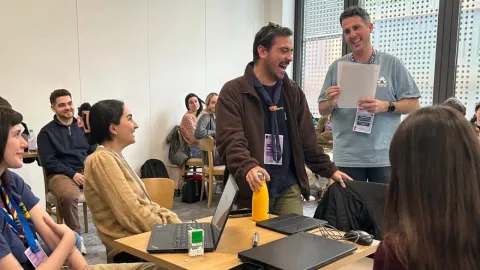 A Portuguese Scout and a facilitator are standing and laughing while other young people around look at them and smile.