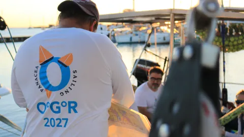 A man wears a t-shirt on a boat. The t-shirt reads Panacea Sailing and OºGºR 2027.