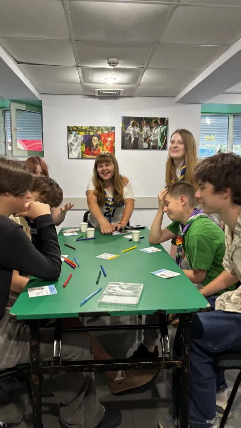 Seven young Ukrainians wearing Scout scarves sit around a table, some of them laughing.