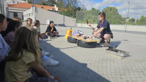 Children sit in a semicircle while looking at a young adult showing objects.