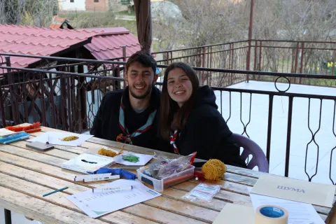 Two young people smile at the camera while sitting in front of a table with drawing materials on it.