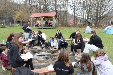 Young people of different ages sit around a campfire while holding papers.