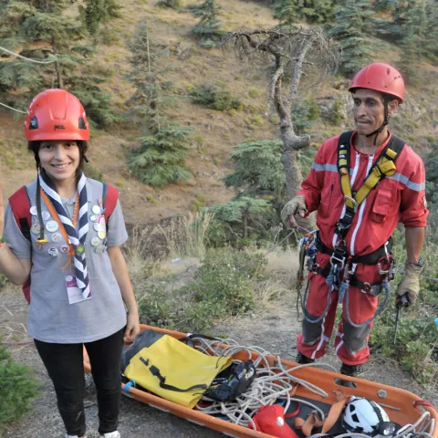 A young woman wears a helmet and makes the Scout symbol with her right hand and a trainer, wearing a helmet and harness, stands next to her and search and rescue equipment during a training session in the Algerian forest