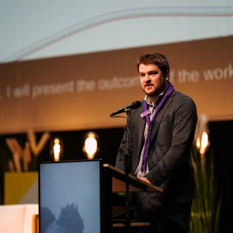 A young person wearing a Scout scarf is speaking on a stage.